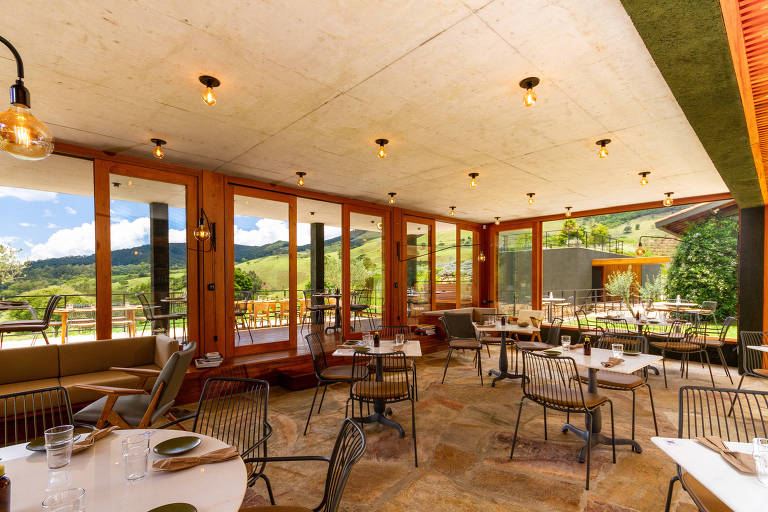 A imagem mostra o interior de um restaurante moderno com grandes janelas de vidro que oferecem uma vista para uma paisagem montanhosa. O espaço é iluminado por lâmpadas pendentes e possui mesas redondas com cadeiras de metal. O piso é de pedras e as paredes são de concreto e madeira, criando um ambiente acolhedor e contemporâneo. Há uma área externa visível através das janelas, com mesas ao ar livre.