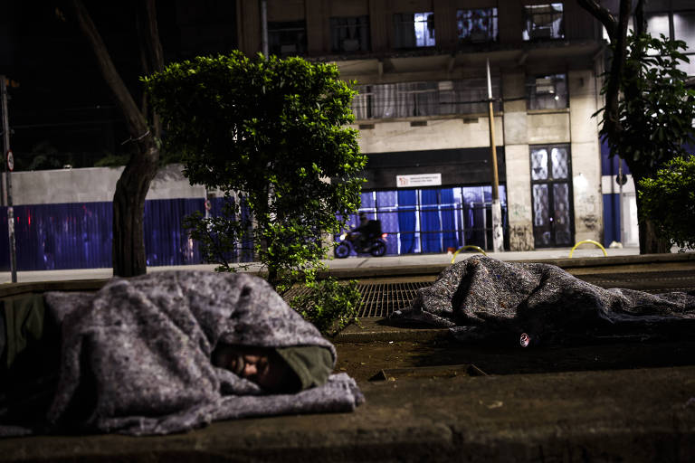 The image shows two people lying on a sidewalk, covered with blankets. They are in an urban setting at night, with buildings in the background and low vegetation to the left. The lighting is dim, and there is a parked motorcycle in the background, as well as an area with blue fences.