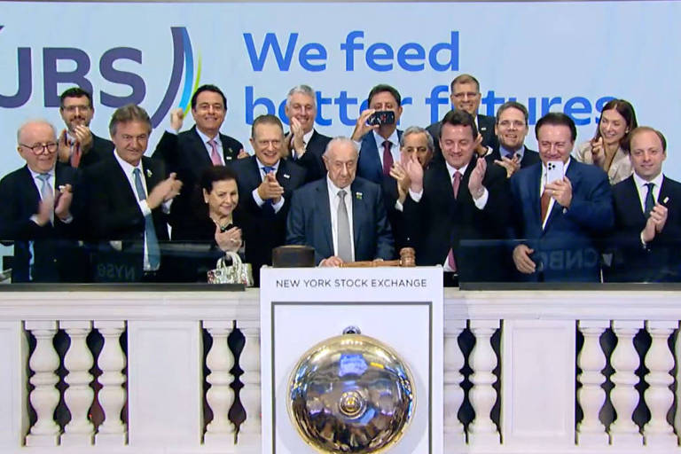 A imagem mostra um grupo de pessoas em uma cerimônia de abertura da Bolsa de Valores de Nova York. Eles estão em frente a um painel com o logotipo da JBS e a frase 'We feed better futures'. As pessoas estão aplaudindo e algumas estão sorrindo, enquanto outras estão em pé ao redor de uma mesa com um sino. O ambiente é festivo e formal, com várias pessoas vestindo ternos.