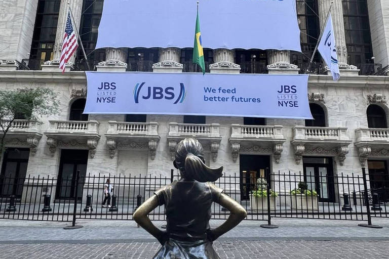 A imagem mostra a famosa estátua da Menina (Fearless Girl) em primeiro plano, de costas, em frente ao edifício da Bolsa de Valores de Nova York. Ao fundo, há grandes banners da empresa JBS, com o texto 'We feed better futures' e o logotipo da empresa. Bandeiras dos Estados Unidos e do Brasil estão visíveis nas laterais. O ambiente é urbano, com a arquitetura clássica do edifício da bolsa.