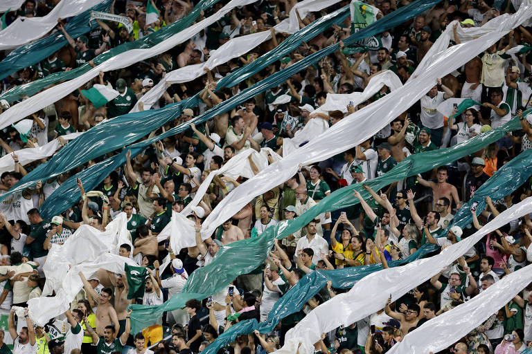 A imagem mostra uma grande multidão de torcedores em um estádio, com muitos deles segurando bandeiras e faixas nas cores verde e branca. A maioria das pessoas está sem camisa, demonstrando entusiasmo e apoio ao time. O ambiente é vibrante, com uma atmosfera de celebração e emoção.