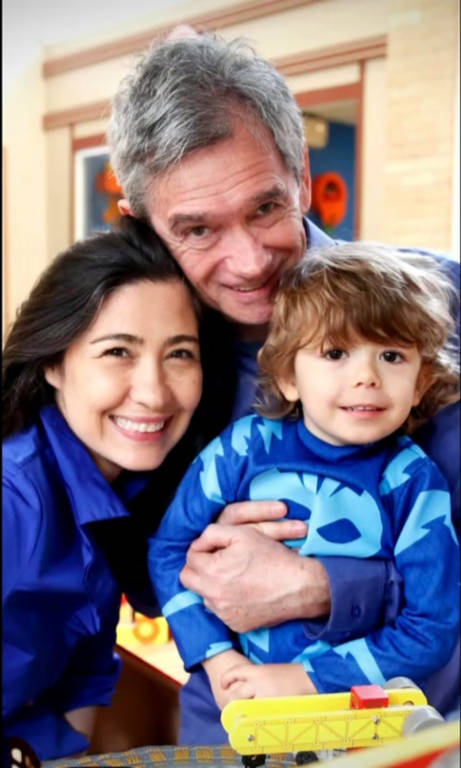 A imagem mostra um homem, uma mulher e uma criança sorrindo juntos. O homem está segurando a criança, que está vestindo uma roupa azul com um desenho. A mulher está ao lado deles, também sorrindo. O fundo parece ser um ambiente interno, possivelmente uma sala de recreação ou uma casa, com cores vibrantes.