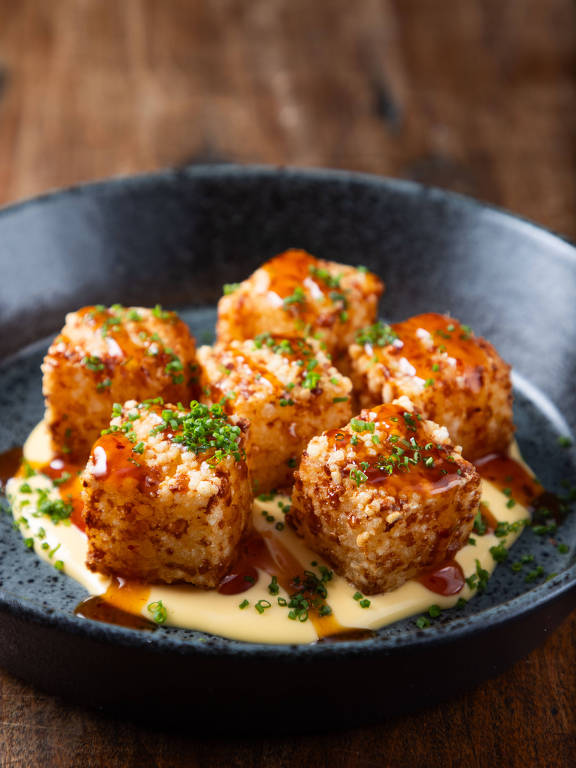 Um prato preto com cinco bolinhos empanados e fritos, cobertos com um molho brilhante e decorados com cebolinha picada. Os bolinhos estão dispostos sobre um molho cremoso claro, com um toque de molho escuro ao redor.