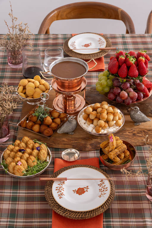 Uma mesa decorada com uma toalha xadrez. No centro, há um recipiente de fondue de chocolate em cobre, cercado por várias travessas com doces. Existem bolinhos fritos, frutas como morangos e uvas, e outros doces dispostos em pratos brancos e de metal. A mesa também possui copos e talheres, criando um ambiente acolhedor e festivo.