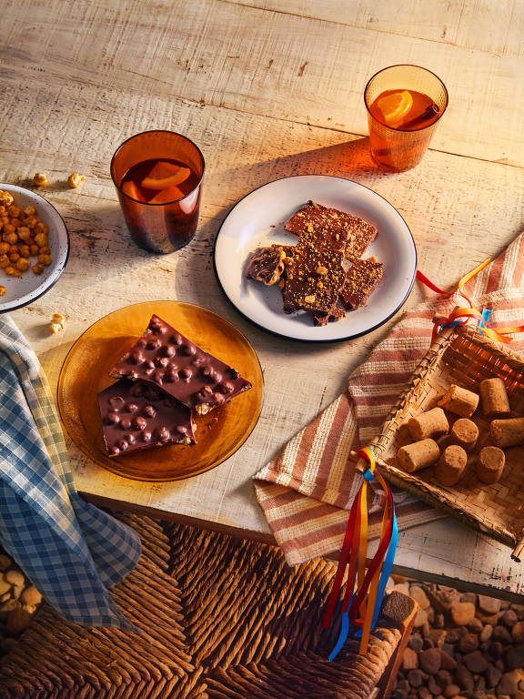 A imagem mostra uma mesa com diversos lanches dispostos. Há um prato branco com doces cobertos de chocolate e granola, um prato amarelo com barras de chocolate, e uma cesta com pequenos doces. Também estão presentes copos laranja com bebida, um pano de mesa xadrez azul e um pano listrado. O fundo é de madeira clara, criando um ambiente acolhedor.