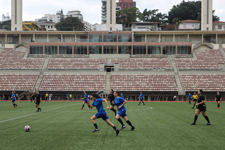 A imagem mostra um jogo de futebol em um estádio. Vários jogadores estão em campo, com alguns vestindo uniformes azuis e outros em uniformes pretos. O campo é coberto por grama sintética e há arquibancadas ao fundo, que estão vazias. Ao fundo, é possível ver prédios e uma área urbana.
