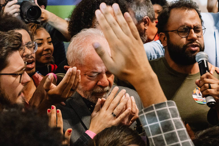A imagem mostra um grupo de pessoas em um evento político, com destaque para um homem de cabelos grisalhos e barba, que parece emocionado, com os olhos fechados e as mãos unidas em um gesto de oração ou agradecimento. Ao seu redor, várias mãos estão levantadas, algumas tocando-o, enquanto outras seguram microfones e câmeras. O ambiente é de intensa interação e apoio emocional.