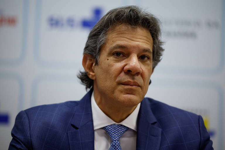 Um homem com cabelo grisalho e liso, usando um terno azul escuro e uma gravata clara. Ele está olhando para a frente com uma expressão séria. O fundo é desfocado, com algumas formas e cores que não são claramente identificáveis.