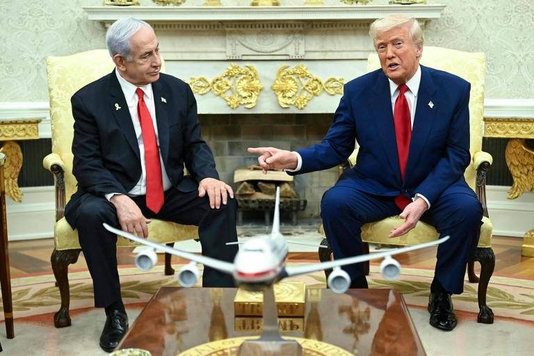 A imagem mostra dois homens sentados em cadeiras em um ambiente formal. À esquerda, um homem com cabelo grisalho e terno escuro, e à direita, um homem com cabelo loiro e terno escuro. Entre eles, há um modelo de avião em uma mesa. O fundo apresenta uma lareira decorada e elementos de luxo.