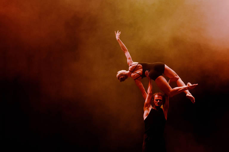 A imagem mostra dois dançarinos em uma apresentação acrobática. Um dançarino, que está em pé, levanta uma dançarina que está em uma posição horizontal, com os braços estendidos. O fundo é nebuloso, com uma mistura de cores quentes, como laranja e vermelho, criando uma atmosfera dramática.