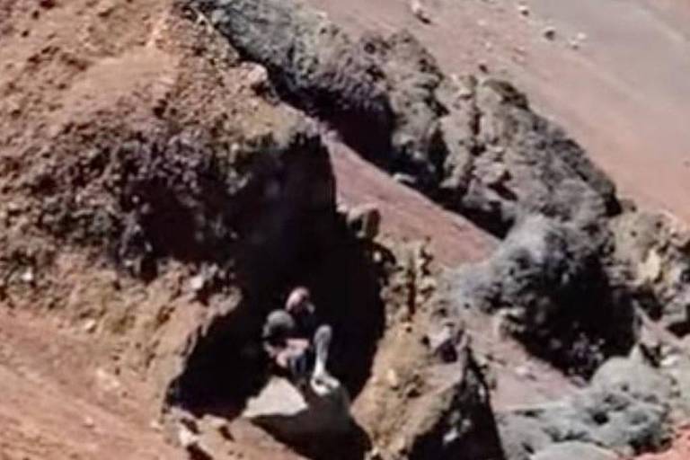 A imagem mostra uma área de terreno vulcânico com formações rochosas irregulares. Duas pessoas estão sentadas em uma das formações, aparentemente explorando o local. O solo é predominantemente marrom e acinzentado, com algumas pedras visíveis ao redor. O ambiente parece árido e desértico, com uma colina ao fundo.