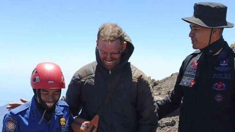 A imagem mostra três homens em um ambiente montanhoso. Um deles, no centro, é um homem de pele clara, com cabelo curto e barba, vestindo uma jaqueta escura e sorrindo. À sua esquerda, um homem com um capacete vermelho e uniforme azul sorri, enquanto à direita, um homem vestido com uma camisa preta e um chapéu também sorri. O fundo é de um céu claro e uma paisagem montanhosa.