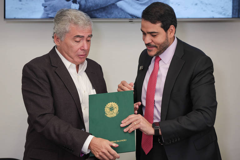 Na imagem, duas pessoas estão em um ambiente interno, participando de uma cerimônia de assinatura. À esquerda, um homem de cabelo grisalho e vestindo uma jaqueta escura e camisa clara, parece estar recebendo um documento de um homem à direita, que usa um terno escuro e gravata. O documento é verde e possui um brasão na capa. Ao fundo, há uma tela com uma imagem em preto e branco.