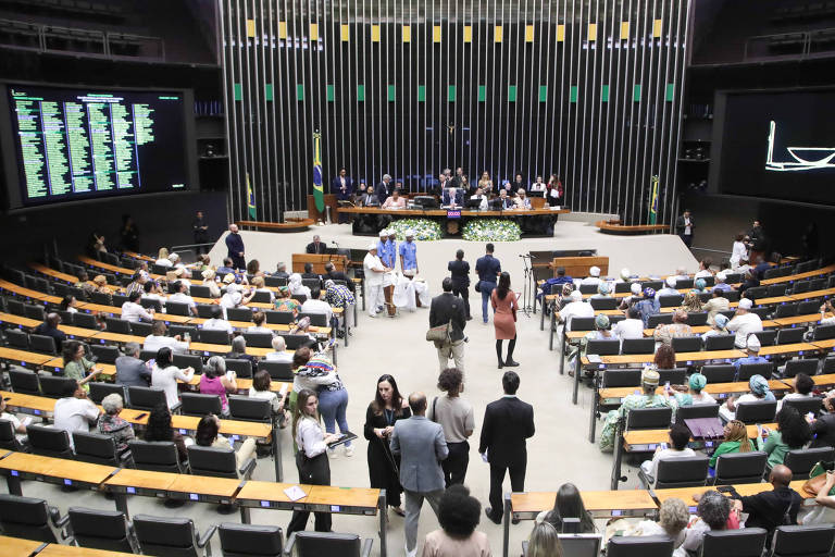 A imagem mostra o interior da Câmara dos Deputados do Brasil, com várias pessoas sentadas nas cadeiras. No fundo, há uma mesa com deputados e bandeiras do Brasil. À esquerda, uma tela exibe uma lista de nomes, enquanto à direita, há uma projeção de um barco. O ambiente é bem iluminado e moderno, com um design arquitetônico contemporâneo.