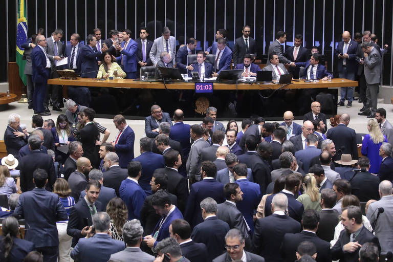 A imagem mostra uma sessão na Câmara dos Deputados do Brasil, com muitos deputados em pé e sentados ao redor de uma mesa central. Há uma bandeira do Brasil ao fundo. O ambiente é formal, com pessoas vestindo trajes sociais. Um cronômetro digital está visível, indicando 01:03. A cena é movimentada, com deputados conversando entre si.