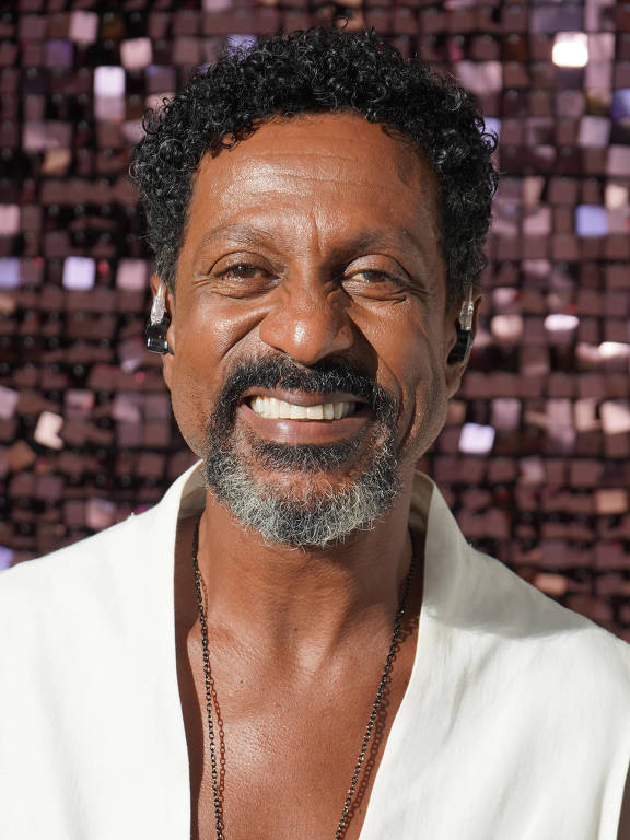 Um homem sorridente com cabelo crespo e barba, vestindo uma blusa sem mangas branca. Ele usa um colar com um pingente vermelho. O fundo é composto por pequenos quadrados brilhantes que refletem a luz, criando um efeito visual interessante.