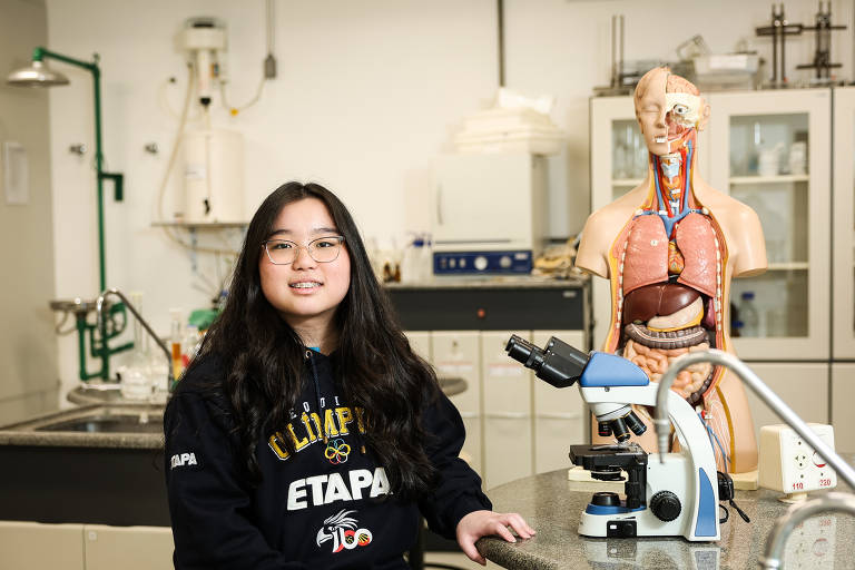 Uma jovem estudante está sentada em uma mesa de laboratório. Ela usa um moletom preto com a inscrição 'ETAPA' e óculos. Ao lado dela, há um microscópio e um modelo anatômico humano em pé, mostrando a estrutura interna do corpo. O fundo do laboratório é composto por armários e equipamentos de ciência.