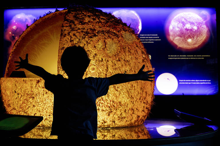 A imagem mostra a silhueta de uma criança com os braços abertos em frente a uma grande representação do Sol, que está iluminada e detalhada. Ao fundo, há painéis informativos sobre o Sol, com imagens e textos visíveis. A cena é predominantemente escura, destacando a figura da criança e a iluminação do modelo do Sol.