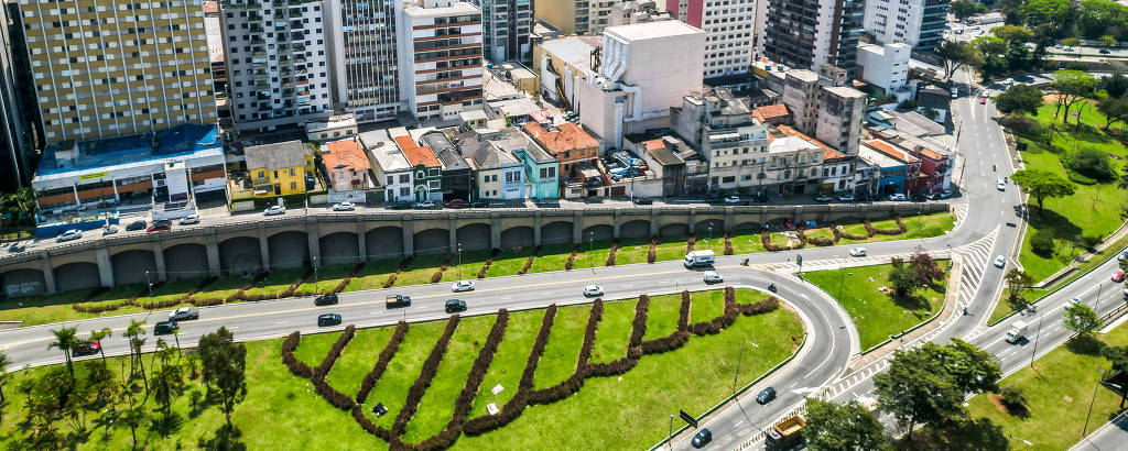 A imagem mostra uma vista aérea de uma área urbana com muitos edifícios altos e modernos. Na parte inferior, há um gramado bem cuidado com canteiros de flores dispostos em um padrão ondulado. Várias ruas e veículos podem ser vistos, além de árvores ao redor. O céu está claro e a cidade parece movimentada.