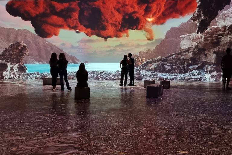 A imagem mostra uma instalação artística onde várias pessoas estão em um espaço amplo, observando uma projeção de uma paisagem dramática. Ao fundo, há uma cena de uma costa com rochas e um céu avermelhado, possivelmente representando uma erupção vulcânica. As pessoas estão em pé ou sentadas em caixas, criando silhuetas contra a luz da projeção.