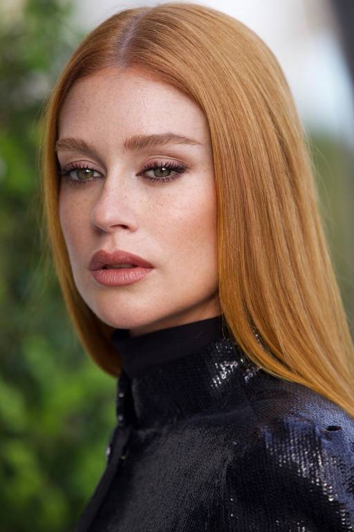 A imagem mostra uma mulher com cabelo liso e longo, de cor ruiva. Ela está usando uma blusa preta brilhante e tem uma expressão séria. O fundo é desfocado, com algumas plantas verdes visíveis.