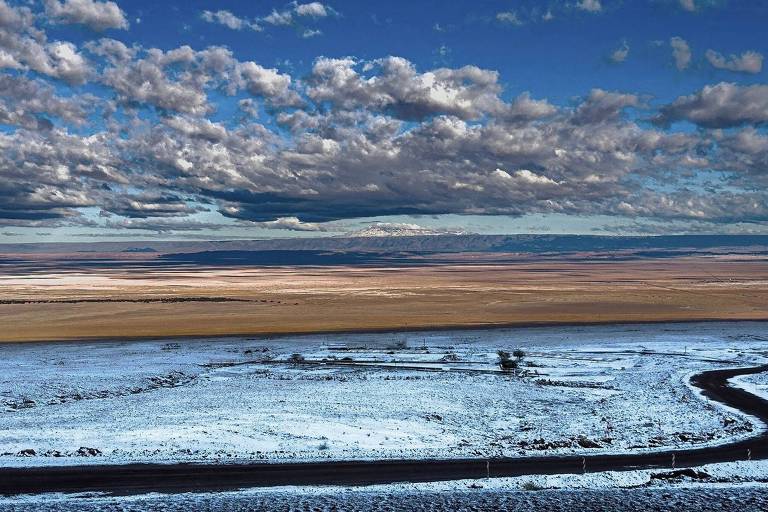 A imagem mostra uma vasta paisagem desértica sob um céu cheio de nuvens. O solo é predominantemente árido, com algumas áreas cobertas de neve ou gelo. No primeiro plano, há uma estrada sinuosa que se estende pela paisagem, enquanto ao fundo, as montanhas são visíveis sob um céu azul com nuvens brancas e cinzas.