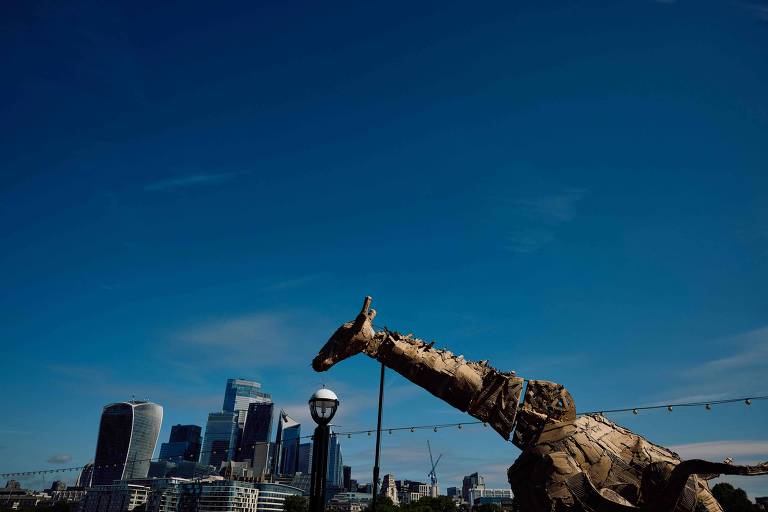 A imagem mostra uma escultura de girafa feita de papelão, posicionada em primeiro plano. Ao fundo, há um skyline urbano com edifícios modernos sob um céu azul claro. A girafa parece estar olhando para a cidade, e há uma lâmpada de rua próxima a ela.
