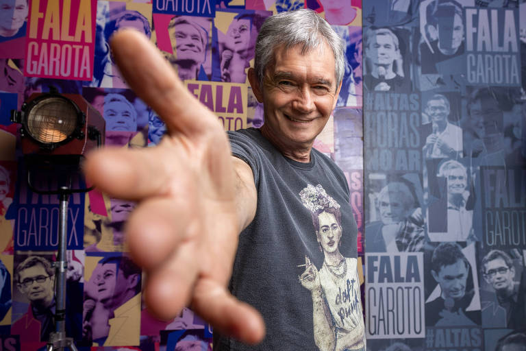 Um homem de cabelos grisalhos, usando uma camiseta preta com uma estampa de uma mulher, está posando para a foto com um gesto de saudação, estendendo a mão em direção à câmera. Ao fundo, há uma parede decorada com várias imagens e textos, incluindo as palavras 'Fala Garoto' e 'Faltas Horas'.