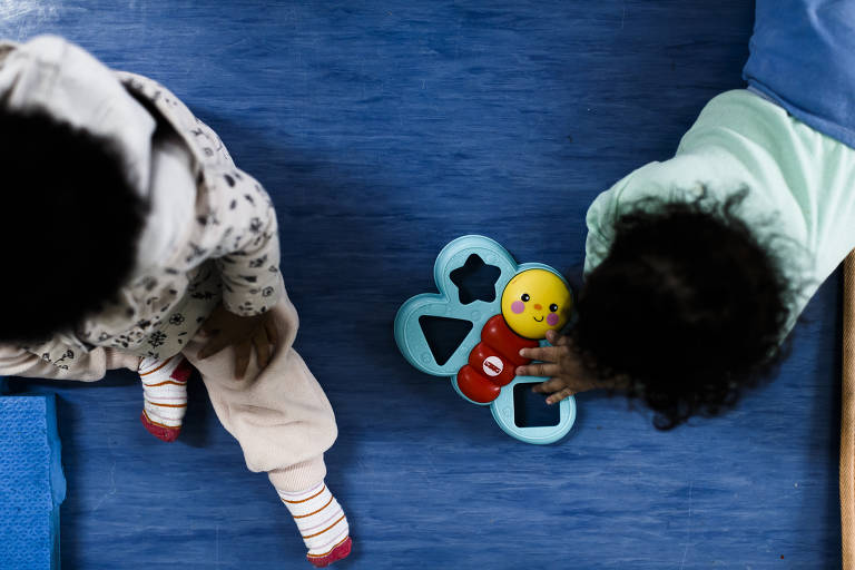 A imagem mostra duas crianças pequenas brincando em um tapete azul. Uma criança está usando uma roupa clara e a outra está vestindo uma roupa com padrões. No centro da imagem, há um brinquedo colorido em forma de inseto, com uma cabeça amarela e corpo vermelho, que uma das crianças está segurando.