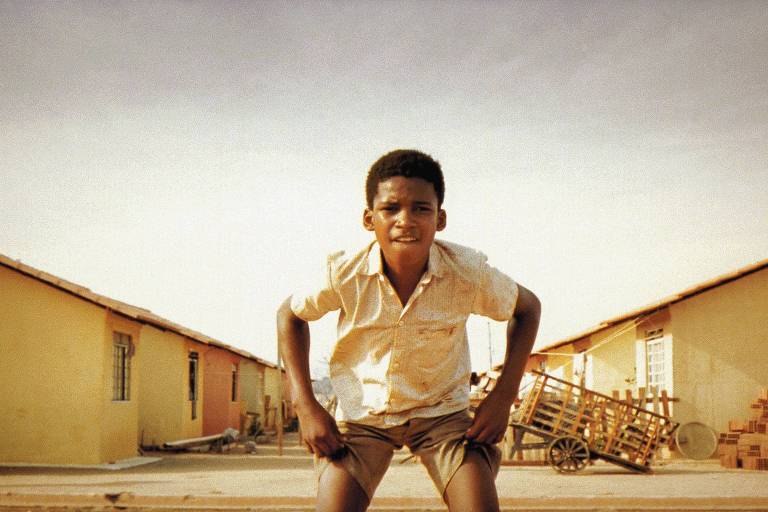 A imagem mostra um menino em um ambiente urbano, posicionado em primeiro plano. Ele está em uma posição de agachamento, com as mãos nos joelhos, olhando para a câmera. Ao fundo, há casas de cor amarela e uma estrutura de madeira, possivelmente um carrinho ou uma carroça, além de alguns tijolos empilhados. O céu está claro, indicando um dia ensolarado.