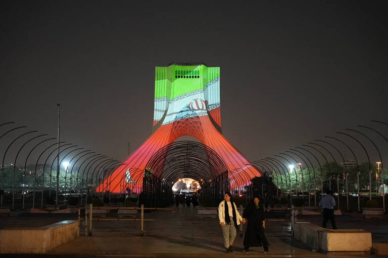 A imagem mostra a Torre Azadi, em Teerã, iluminada com as cores da bandeira do Irã, verde, branco e vermelho. A torre é projetada com um padrão que inclui a mão segurando uma bandeira. No primeiro plano, duas pessoas caminham sob a estrutura, enquanto o ambiente ao redor é noturno, com iluminação visível e árvores ao fundo.