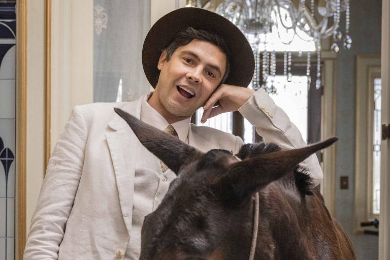 Um homem vestido com um terno claro e chapéu está posando ao lado de um burro em um ambiente interno. O homem sorri e parece estar se divertindo, enquanto o burro está com a cabeça baixa, próximo ao homem. O fundo apresenta uma decoração elegante, com um lustre visível e paredes decoradas.
