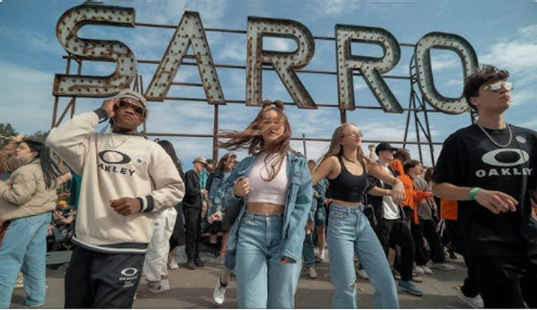 A imagem mostra um grupo de jovens dançando em frente a um grande letreiro que diz 'SARRO'. Os dançarinos estão vestidos de forma casual, com roupas de marca, e alguns usam óculos escuros. O ambiente parece ser ao ar livre, com várias pessoas ao fundo, algumas observando e outras participando da dança. O céu está parcialmente nublado.