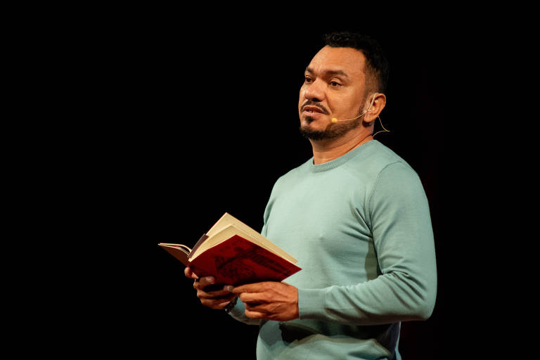 Um homem está em um palco, segurando um livro aberto. Ele usa uma camiseta de manga longa azul clara e tem um microfone preso à orelha. O fundo é escuro, destacando sua figura enquanto ele parece estar falando ou apresentando algo.