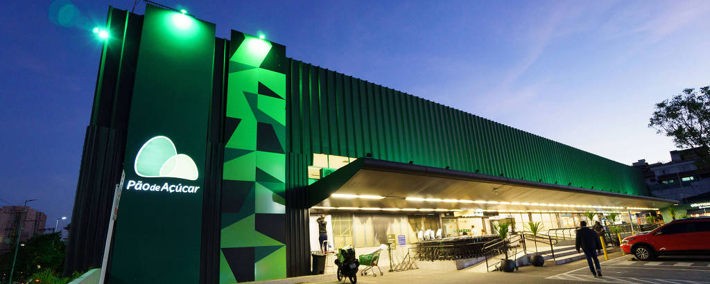 A imagem mostra a fachada de uma loja Pão de Açúcar ao entardecer. O edifício é moderno, com uma estrutura metálica e iluminação verde. Há vagas de estacionamento na frente, algumas delas reservadas para pessoas com deficiência. O ambiente é urbano, com árvores ao fundo e um carro vermelho estacionado.