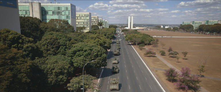 A imagem mostra uma rua em Brasília, cercada por árvores, com uma fila de veículos militares se deslocando. Ao fundo, é possível ver edifícios modernos e o famoso Congresso Nacional, com suas cúpulas distintas. O céu está parcialmente nublado, e a cena parece ser de um evento militar ou desfile.