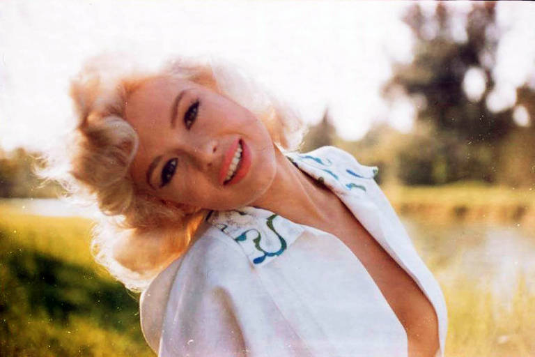 A imagem mostra uma mulher com cabelo loiro e ondulado, sorrindo para a câmera. Ela está vestindo uma blusa branca com detalhes coloridos e está em um ambiente ao ar livre, com grama verde e árvores ao fundo. A luz do sol ilumina a cena, criando um efeito suave.