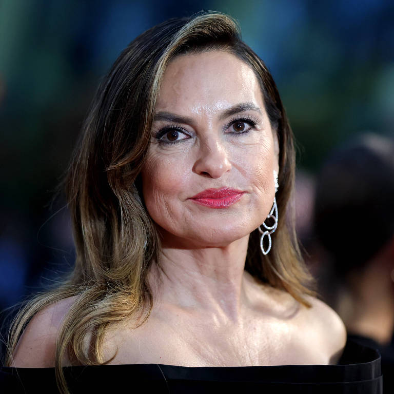 Uma mulher com cabelo longo e liso, usando um vestido preto sem mangas. Ela tem brincos elegantes e um batom vermelho. O fundo é desfocado, sugerindo que a imagem foi tirada em um evento social ou tapete vermelho.