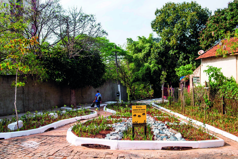 A imagem mostra um espaço urbano ajardinado com caminhos de pedras e canteiros de plantas. À esquerda, há uma pessoa caminhando em direção a um caminho que se curva. No centro, há uma placa amarela com texto visível, e ao fundo, árvores e uma parede de concreto. O ambiente é ensolarado e bem cuidado, com vegetação ao redor.