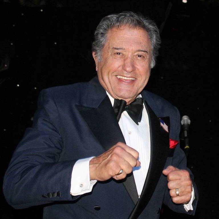 Um homem sorridente está usando um traje formal, composto por um paletó escuro e uma gravata borboleta. Ele parece estar se divertindo, com as mãos levantadas em um gesto de celebração. O fundo é escuro, possivelmente um palco ou um evento noturno, com luzes brilhantes ao fundo.