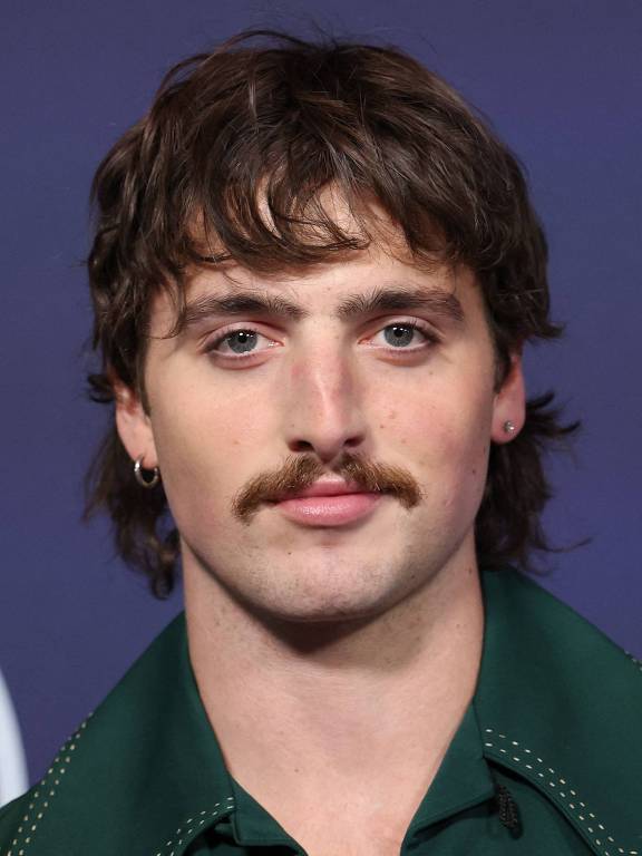 Um homem com cabelo castanho e ondulado, usando uma camisa verde. Ele tem um bigode e está olhando diretamente para a câmera. O fundo é de cor roxa com um logotipo que diz 'AMERICAN MUSIC'.