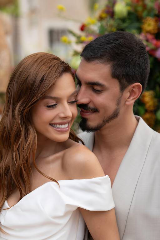 Um casal sorridente está posando para a foto em um ambiente decorado com flores. A mulher tem cabelo longo e castanho claro, vestindo um vestido branco com um decote ombro a ombro. O homem, com cabelo curto e barba bem aparada, está próximo dela, sorrindo e olhando para ela com carinho. O fundo é composto por uma variedade de flores coloridas, criando uma atmosfera romântica.
