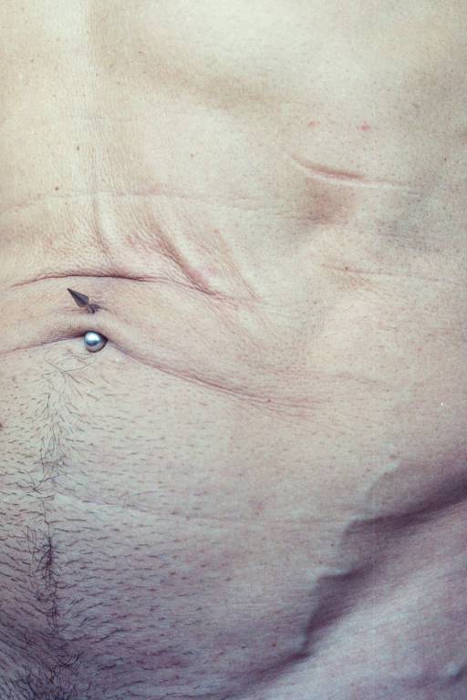 A imagem mostra um close do abdômen de uma pessoa, com pele visivelmente enrugada e marcas. Há um piercing visível na região do umbigo, e a textura da pele é destacada, mostrando pelos e irregularidades.