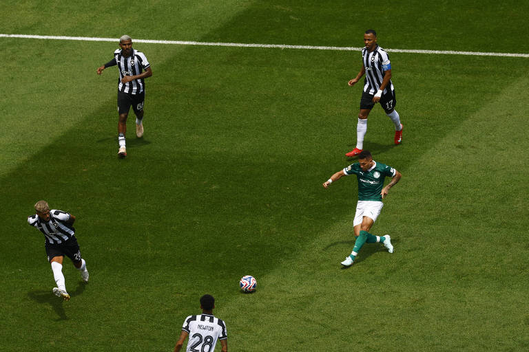 A imagem mostra um jogo de futebol em campo. Um jogador vestido com uma camisa verde e calças brancas está correndo com a bola em direção ao gol, enquanto quatro jogadores adversários, todos vestidos com camisas listradas em preto e branco, estão se posicionando para tentar interceptá-lo. O campo é bem iluminado e a grama está verde e bem cuidada.