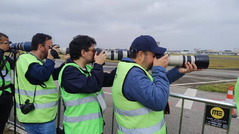 Um grupo de fotógrafos está posicionado em uma área de observação, todos usando coletes verdes. Eles estão segurando câmeras com lentes longas, apontando-as para o horizonte. O ambiente parece ser um aeroporto, com uma pista visível ao fundo e um céu nublado.