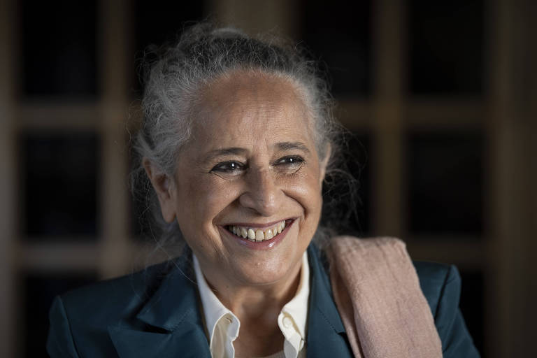 A imagem mostra uma mulher sorridente com cabelo grisalho preso em um coque. Ela está vestindo um paletó azul e uma camisa branca, com um lenço ou xale rosa sobre o ombro. O fundo é desfocado, destacando seu rosto iluminado e expressivo.