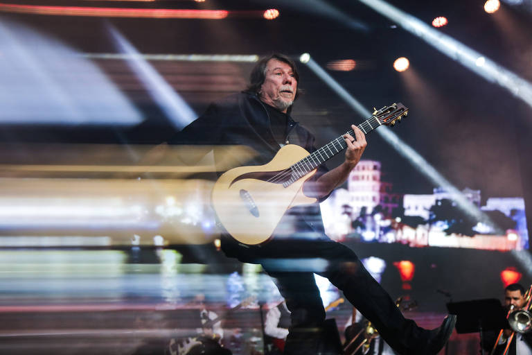 Um músico está se apresentando em um palco, tocando um violão. Ele está em movimento, com uma expressão intensa, e a imagem mostra um efeito de desfoque que sugere velocidade. O fundo é iluminado com luzes coloridas e há uma plateia visível ao fundo, criando uma atmosfera de show ao vivo.