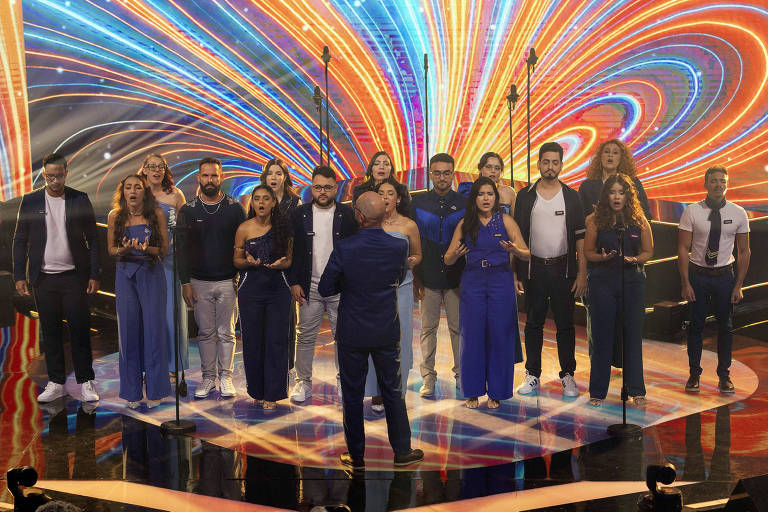 Um grupo de cantores está se apresentando em um palco iluminado com um fundo colorido e vibrante. O grupo é composto por homens e mulheres, todos vestidos de maneira semelhante, com destaque para roupas azuis. Um dos cantores, que está de costas, parece ser o líder ou maestro da apresentação. O ambiente é festivo e cheio de energia, com microfones posicionados para a performance.