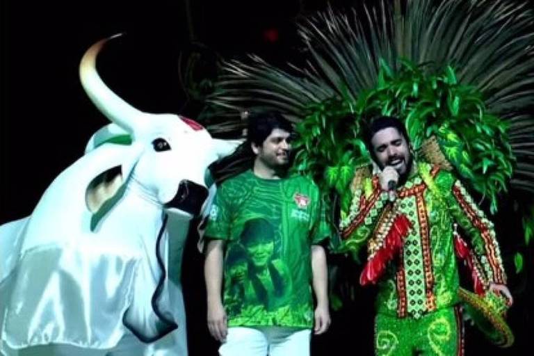 A imagem mostra três pessoas em um palco. À esquerda, uma pessoa vestida com uma fantasia de boi, que é branca com detalhes vermelhos. No centro, uma pessoa usa uma camiseta verde com uma imagem de uma pessoa e calças brancas. À direita, uma pessoa está vestida com um traje colorido, predominantemente verde, com detalhes em vermelho e amarelo, e possui uma grande plumagem na cabeça. O fundo é escuro, destacando os trajes coloridos.
