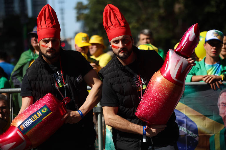 Dois homens estão vestidos com roupas pretas e chapéus vermelhos, segurando objetos infláveis que se assemelham a garrafas de cerveja. Eles estão em um evento ao ar livre, com uma multidão ao fundo, onde pessoas vestem camisetas amarelas e verdes, sugerindo uma celebração ou festa. O ambiente é festivo e animado.